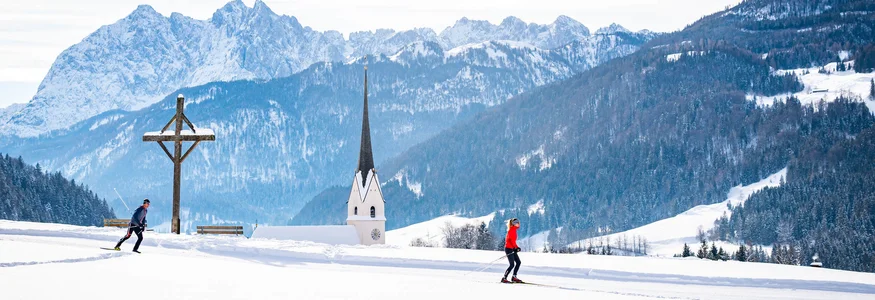 Die Kaiserwinklloipe © Tourismusverband Kaiserwinkl Zwei Langläufer in verschneiter Landschaft mit Kirche und Bergen im Hintergrund