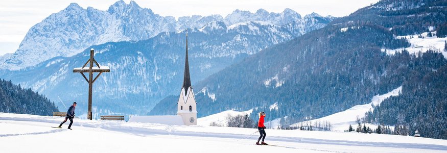 Zwei Langläufer in verschneiter Landschaft mit Kirche und Bergen im Hintergrund