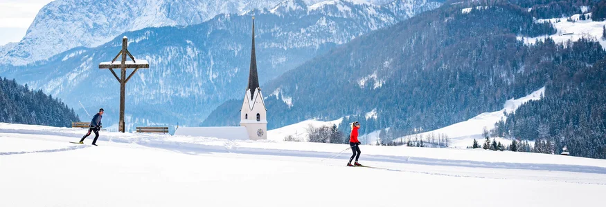 Zwei Langläufer in verschneiter Landschaft mit Kirche und Bergen im Hintergrund
