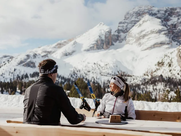 Two people sitting outside with skis in front of snowy mountains