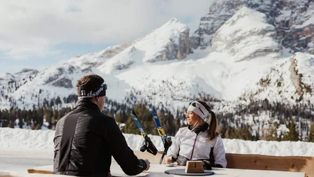Two people sitting outside with skis in front of snowy mountains