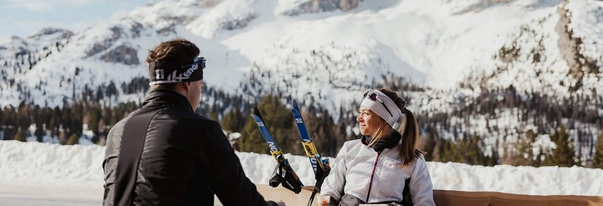 Zwei Personen sitzen draußen mit Skiern vor schneebedeckten Bergen
