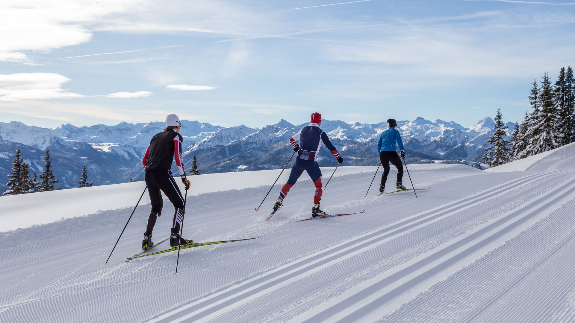 Three cross-country skiers on snowy mountain trail with alpine background