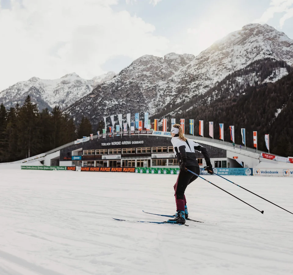 Coop FIS Tour de Ski performance by Le Gruyère AOP © Alex Moling Skilangläufer vor verschneiten Bergen und Nordic Arena in Toblach