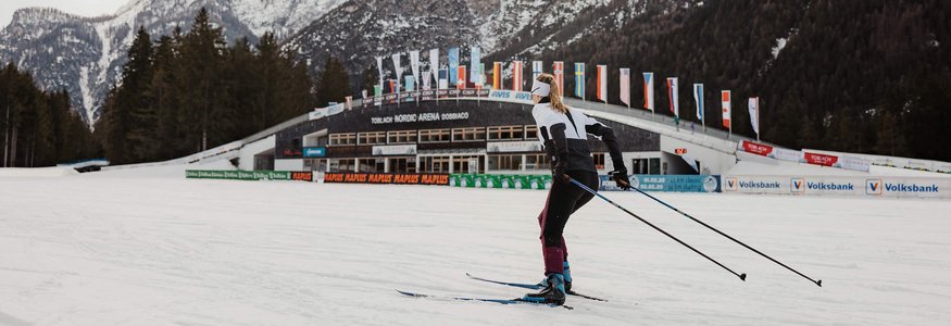 Skilangläufer vor verschneiten Bergen und Nordic Arena in Toblach