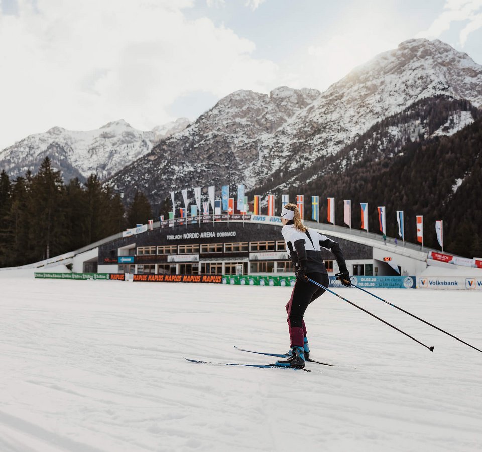 Coop FIS Tour de Ski performance by Le Gruyère AOP © Alex Moling Skilangläufer vor verschneiten Bergen und Nordic Arena in Toblach