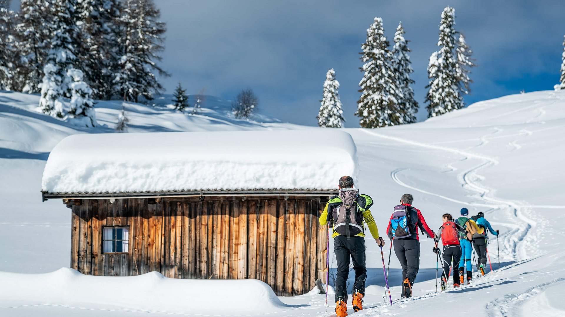 Gruppe Skitourengeher wandert im Schnee an einer verschneiten Holzhütte vorbei