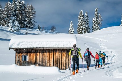 Gruppe Skitourengeher wandert im Schnee an einer verschneiten Holzhütte vorbei