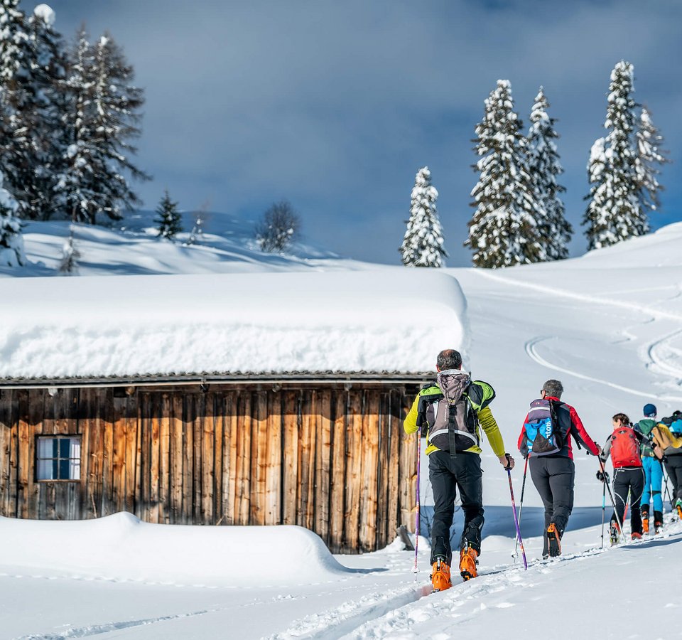 Gruppe Skitourengeher wandert im Schnee an einer verschneiten Holzhütte vorbei