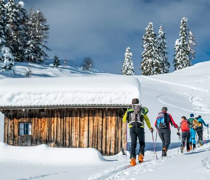 Gruppe Skitourengeher wandert im Schnee an einer verschneiten Holzhütte vorbei