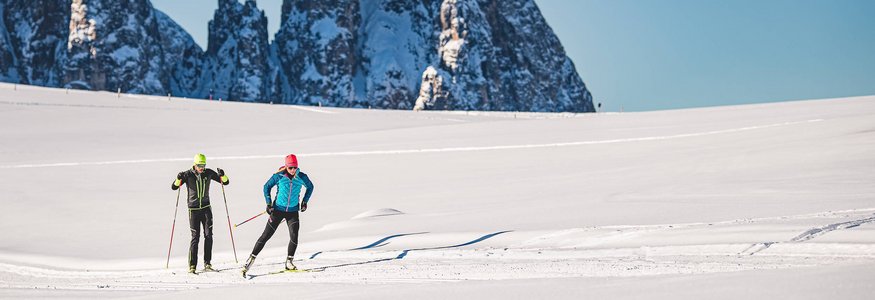 Zwei Langläufer auf schneebedecktem Feld vor hohen, schneebedeckten Bergen