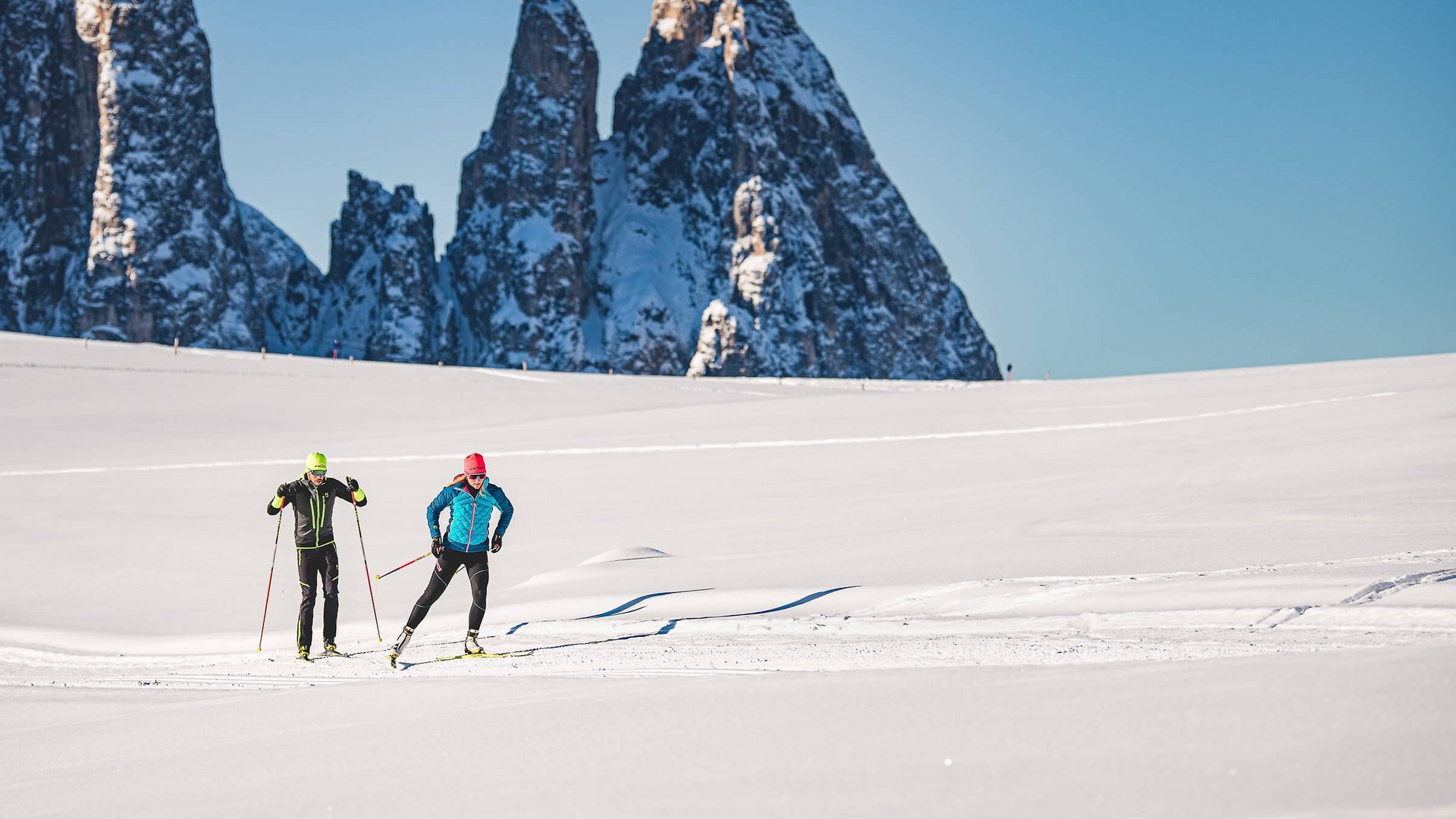 Zwei Langläufer auf schneebedecktem Feld vor hohen, schneebedeckten Bergen