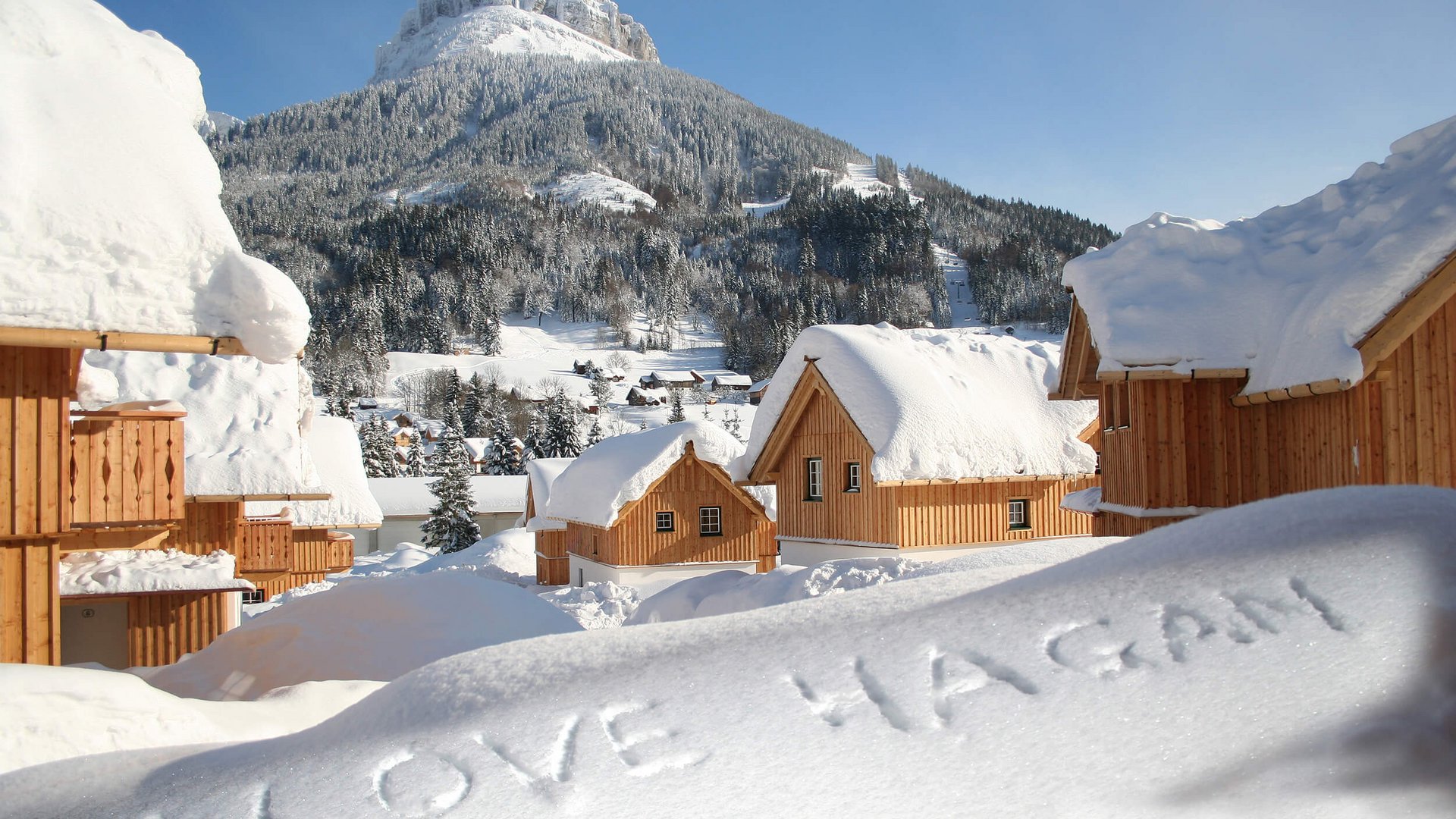 Snow-covered wooden cabins with I LOVE WAGEN written in snow and mountain