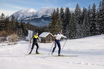 Zwei Langläufer in Winterlandschaft mit schneebedeckten Bäumen und Bergen