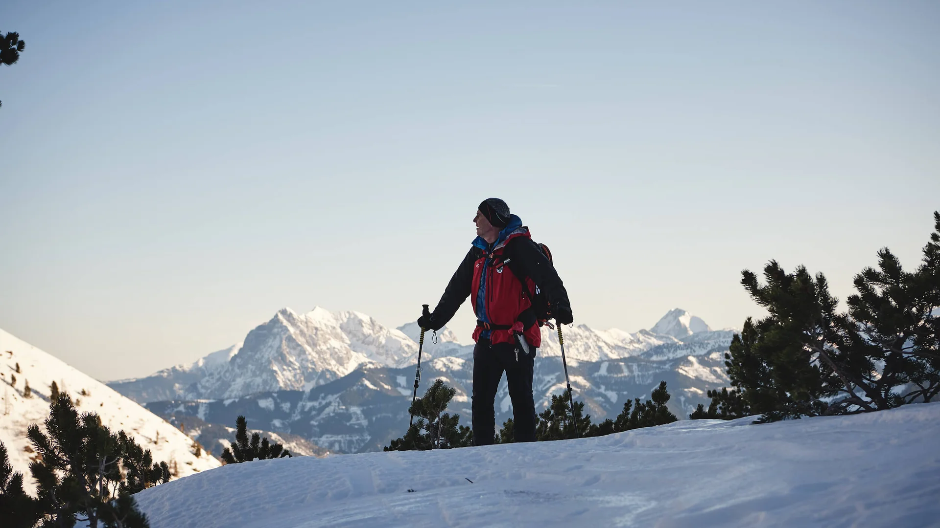 Wanderer mit rotem Rucksack und Stöcken auf schneebedecktem Berg mit Bergkulisse