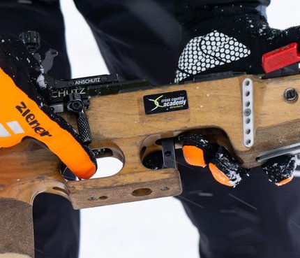 Person hält Biathlon-Gewehr mit orangenen Handschuhen im Schnee