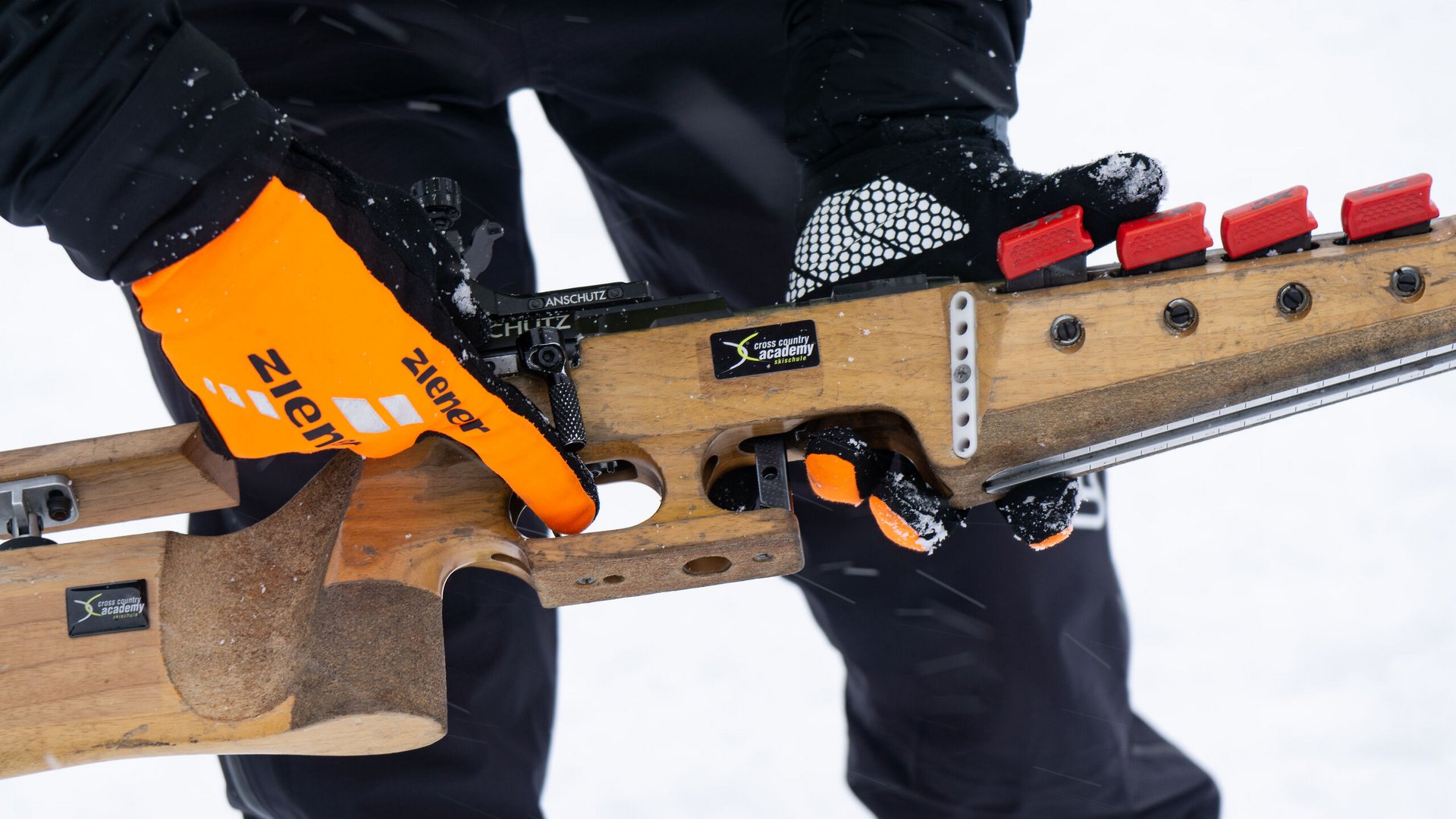Person hält Biathlon-Gewehr mit orangenen Handschuhen im Schnee