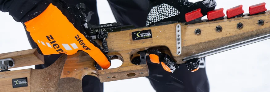 Person hält Biathlon-Gewehr mit orangenen Handschuhen im Schnee