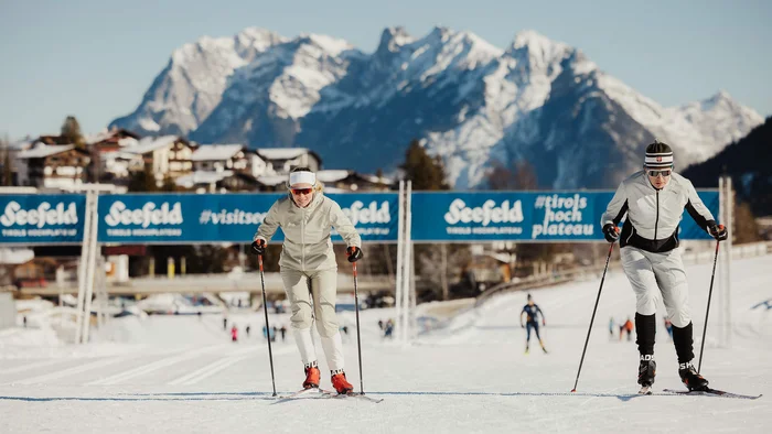 Zwei Langläufer auf der Loipe mit schneebedeckten Bergen im Hintergrund