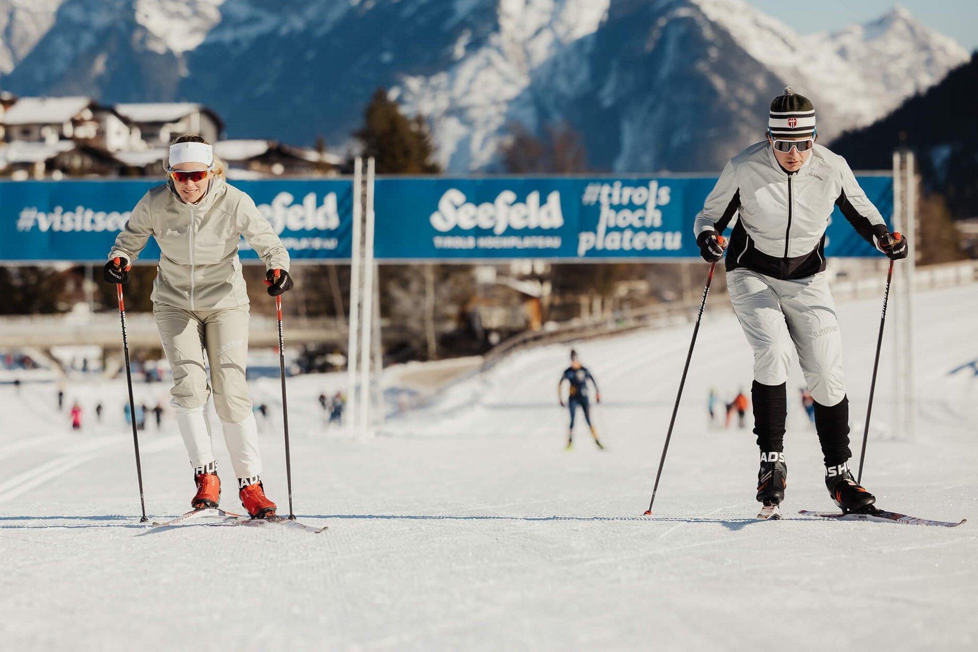 Two cross-country skiers on a trail with snowy mountains in the background