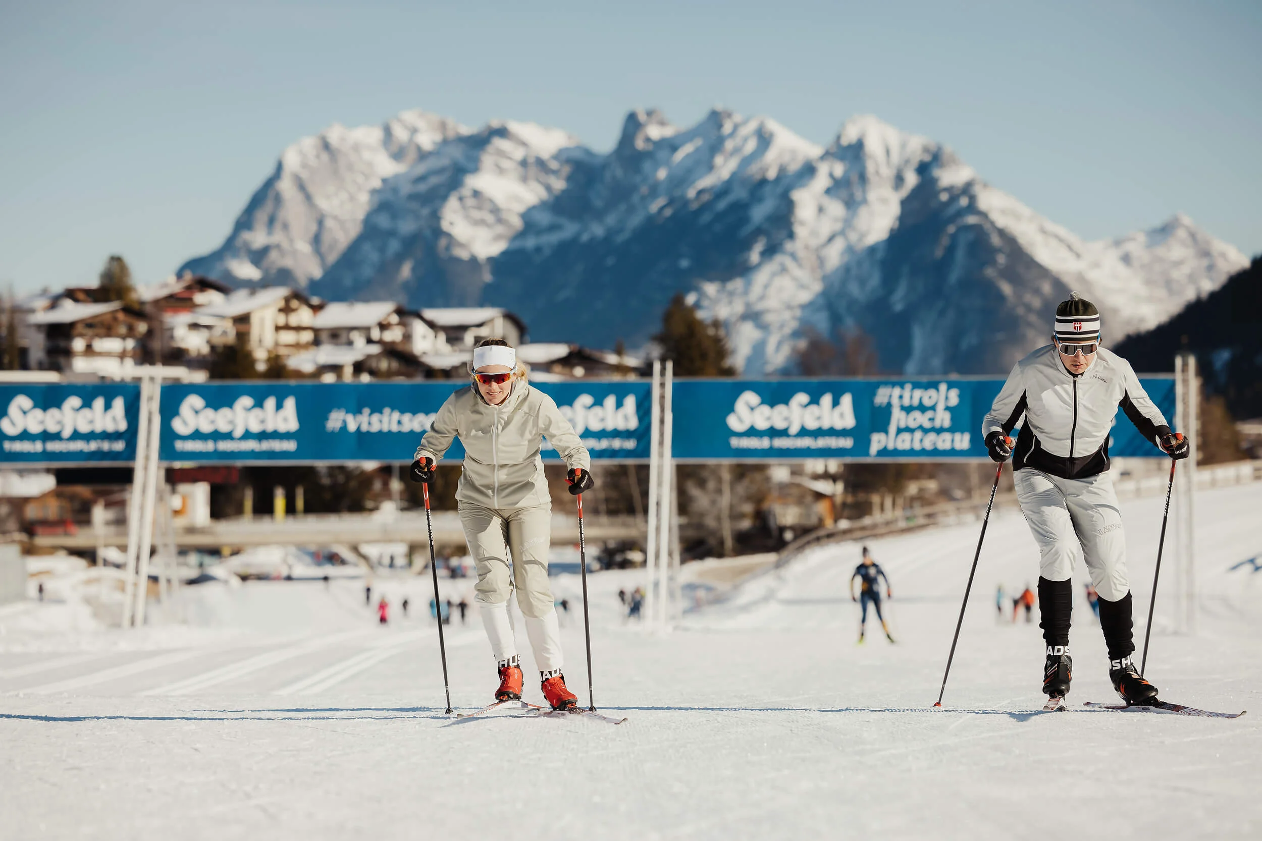 Zwei Langläufer auf der Loipe mit schneebedeckten Bergen im Hintergrund