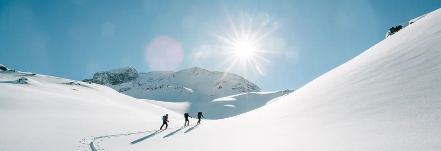 Drei Personen wandern im Schnee bei strahlendem Sonnenschein in den Bergen