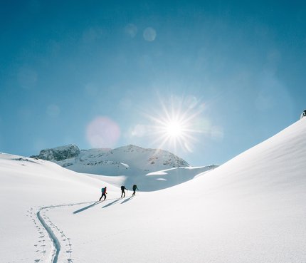 Drei Personen wandern im Schnee bei strahlendem Sonnenschein in den Bergen