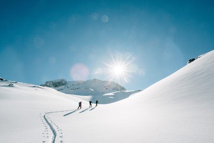 Drei Personen wandern im Schnee bei strahlendem Sonnenschein in den Bergen