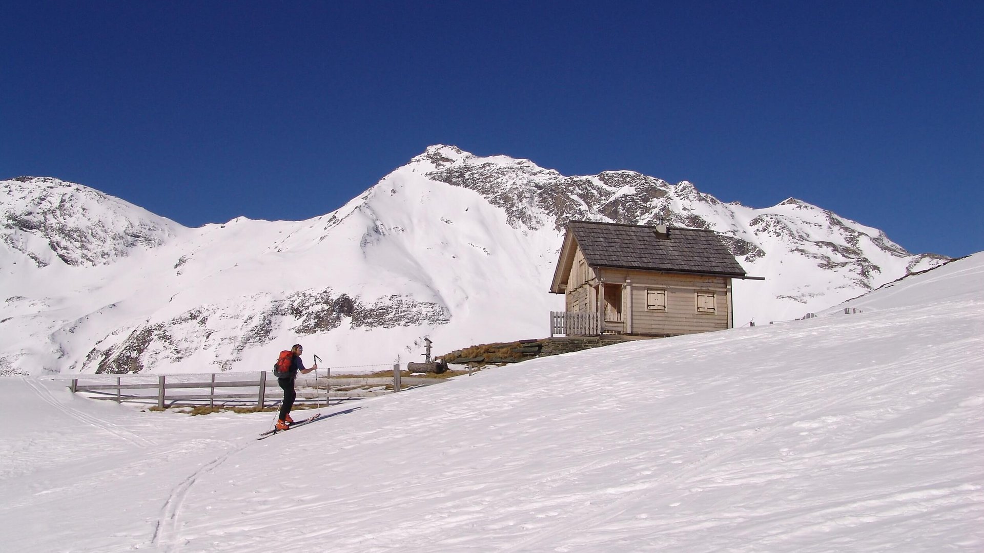 Skier next to a wooden hut in snowy mountain area under clear blue sky