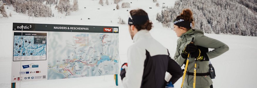 Zwei Skilangläufer schauen auf eine Karte von Nauders und Reschenpass im Schnee