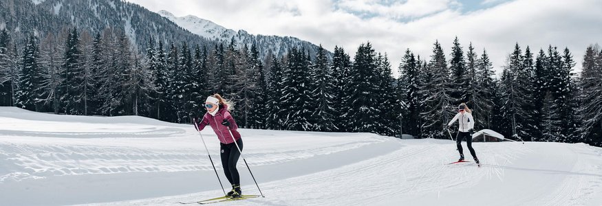 Zwei Frauen beim Langlaufen auf schneebedeckter Strecke mit Bergen und Bäumen