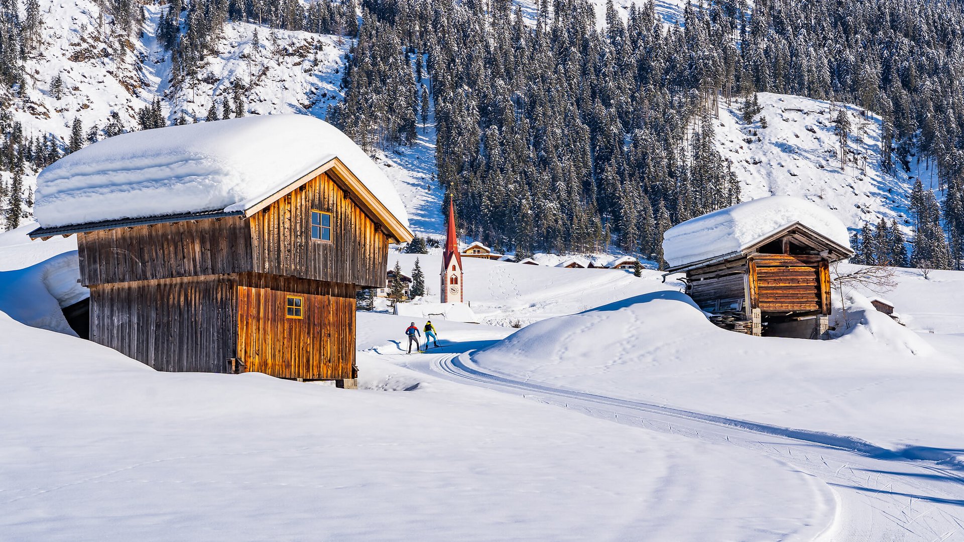 Zwei Langläufer auf verschneiter Spur zwischen Holzscheunen im Winter