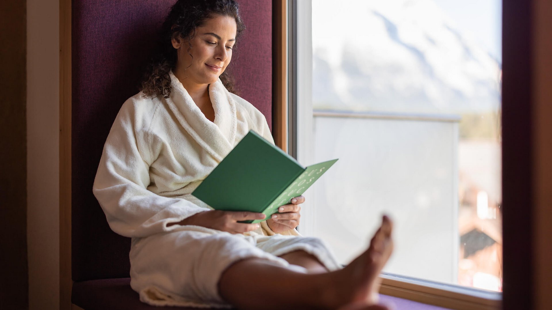 Woman in bathrobe reading book by window with mountain view