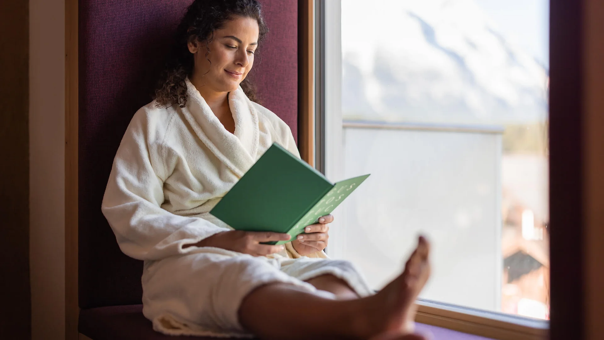 Frau im Bademantel liest entspannt Buch am Fenster mit Bergblick