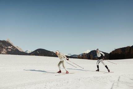 Zwei Personen beim Langlaufen auf schneebedecktem Feld vor Bergen