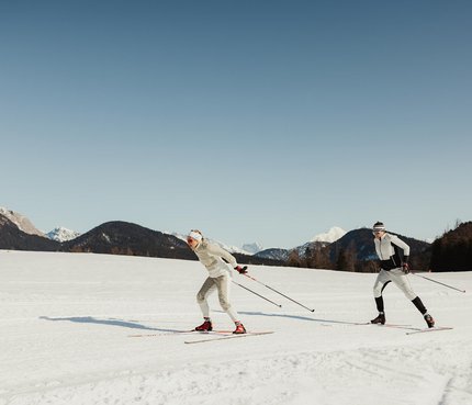 Zwei Personen beim Langlaufen auf schneebedecktem Feld vor Bergen