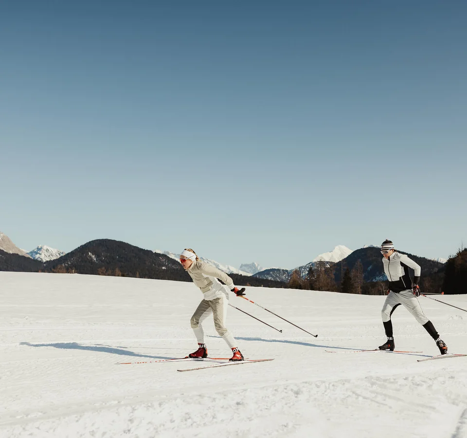 Zwei Personen beim Langlaufen auf schneebedecktem Feld vor Bergen