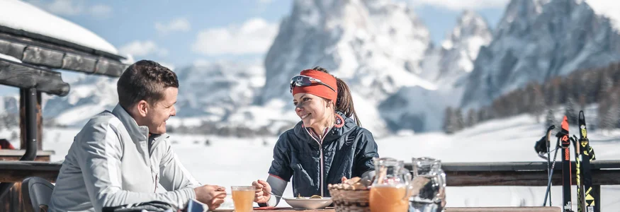 Langlauf & Kultur vereint auf der Seiser Alm © Dolomiti Nordic Ski - Manuel Kottersteger Paar beim Frühstück im Freien mit Schneelandschaft und Bergen im Hintergrund