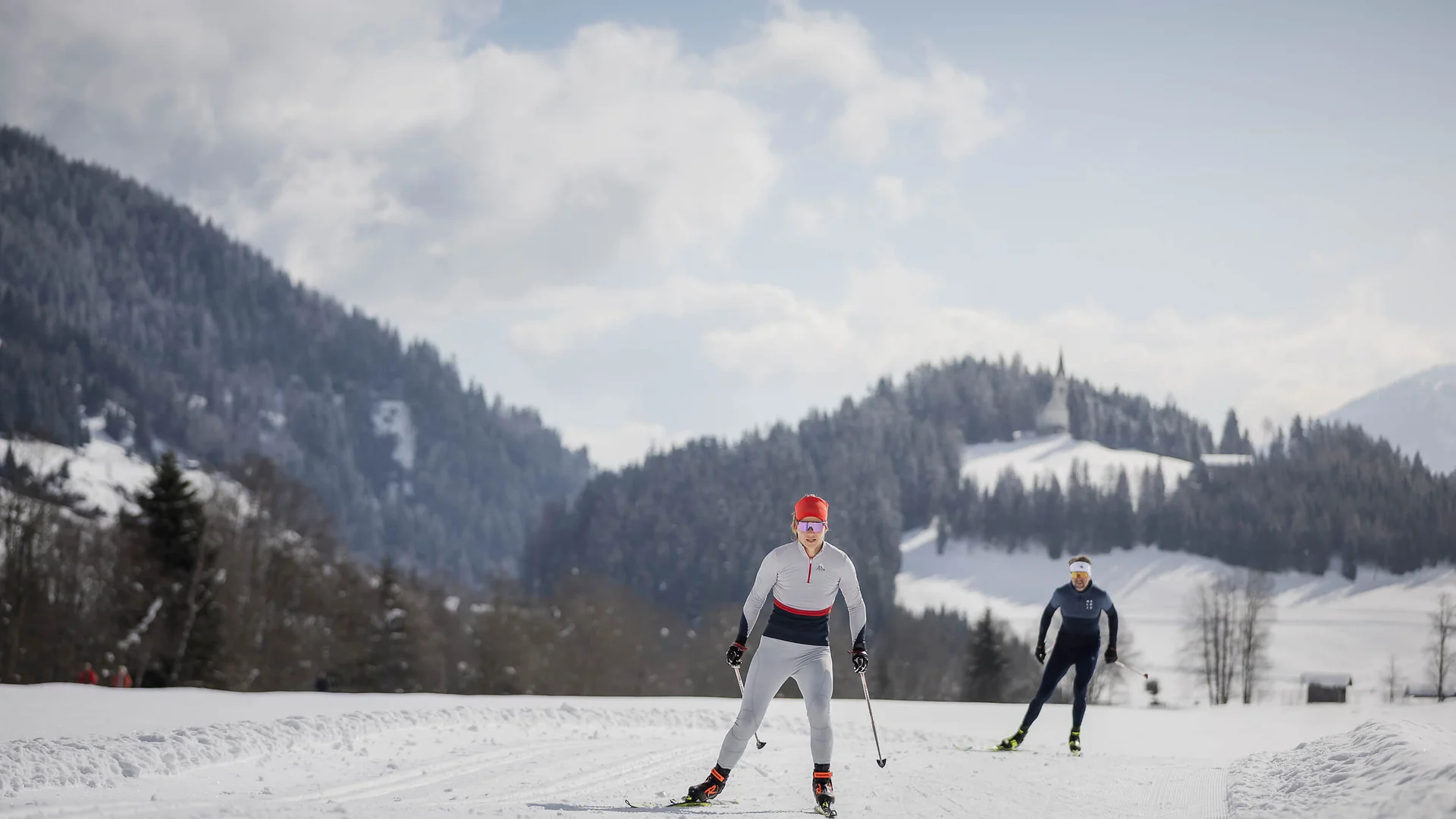 Zwei Langläufer auf verschneiter Loipe vor bewaldeten Bergen