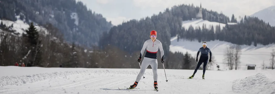Zwei Langläufer auf verschneiter Loipe vor bewaldeten Bergen