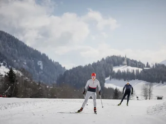 Two cross-country skiers on snowy trail with forested mountains
