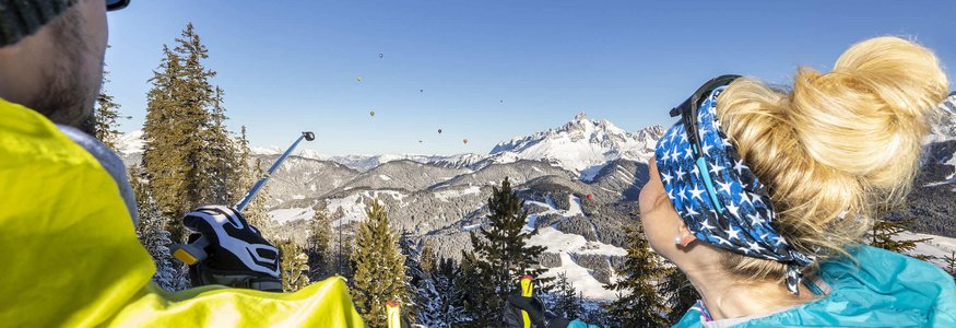 Zwei Wanderer blicken auf schneebedeckte Berge und Heißluftballons im Himmel