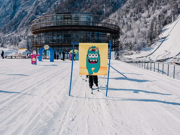 Kind auf Ski hinter einem bunten Monster-Schild vor einem runden Gebäude im Schnee