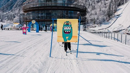 Kind auf Ski hinter einem bunten Monster-Schild vor einem runden Gebäude im Schnee