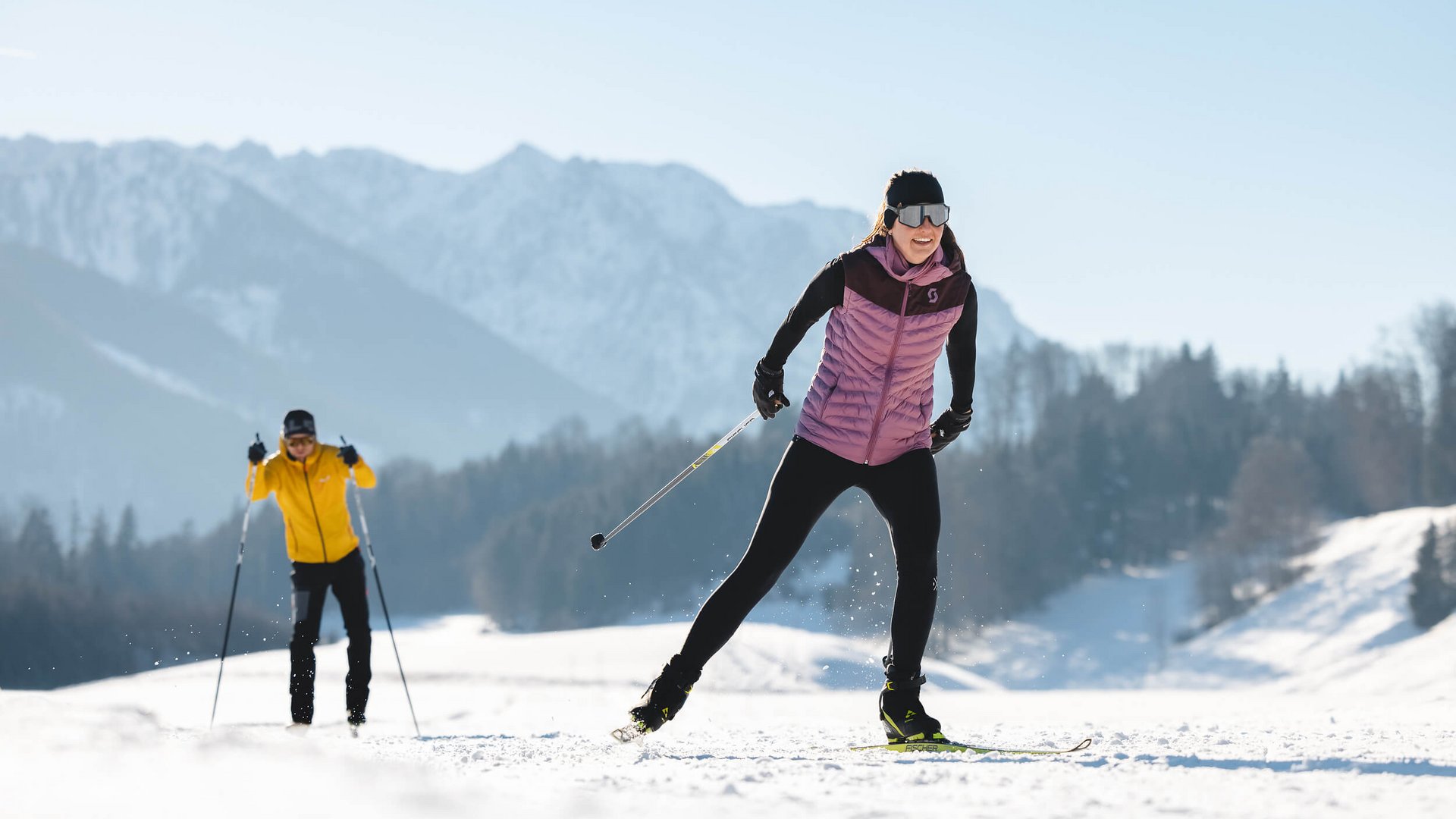 Zwei Personen beim Langlaufen vor verschneiten Bergen.