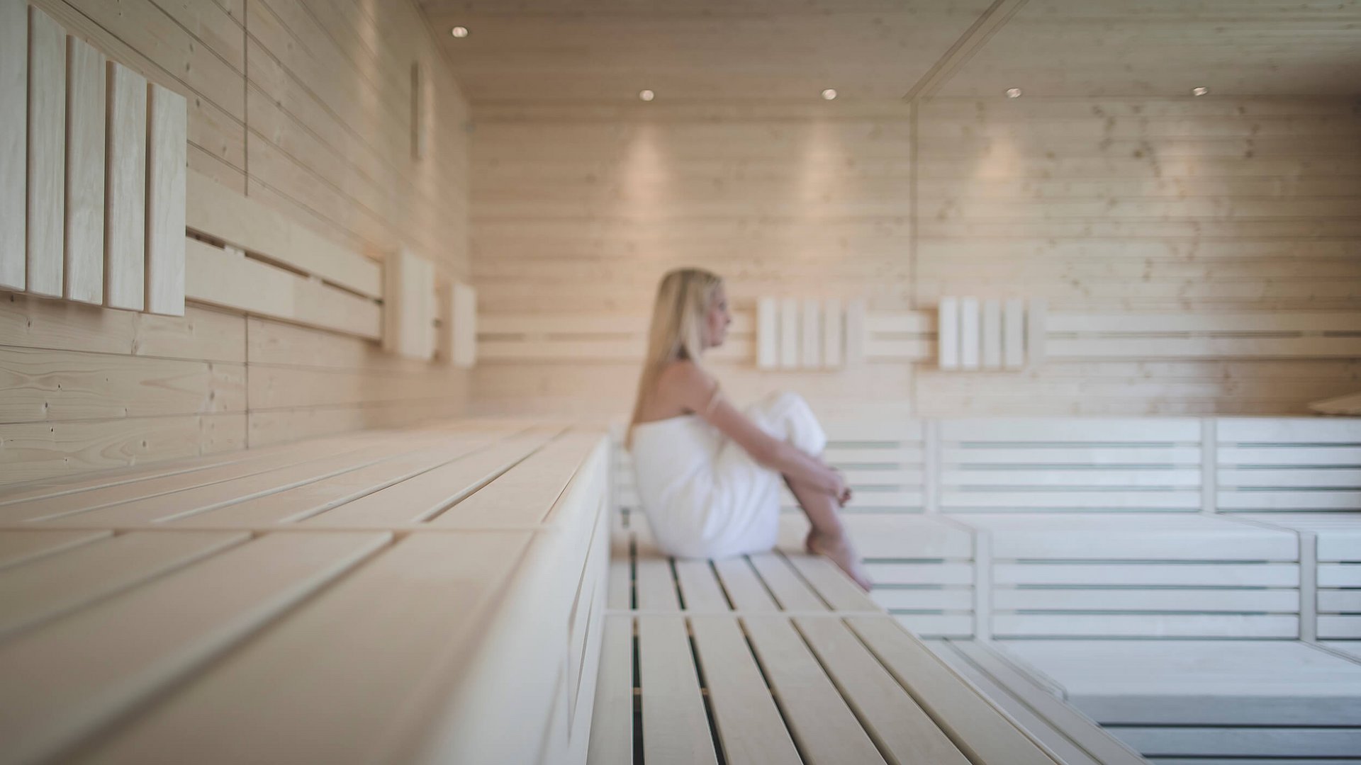 Frau sitzt auf Holzbänken in heller Sauna mit Handtuch
