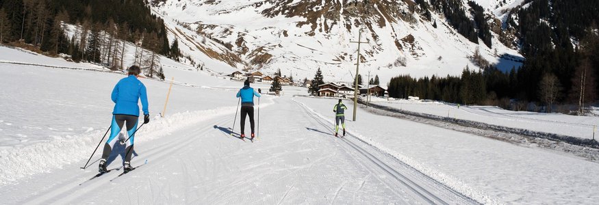 Drei Langläufer fahren auf verschneiter Loipe in den Bergen bei klarem Himmel