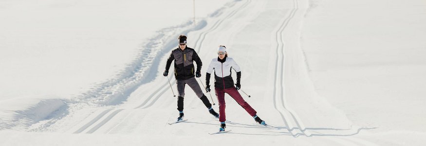 Zwei Personen beim Langlaufen auf einer verschneiten Loipe in den Bergen