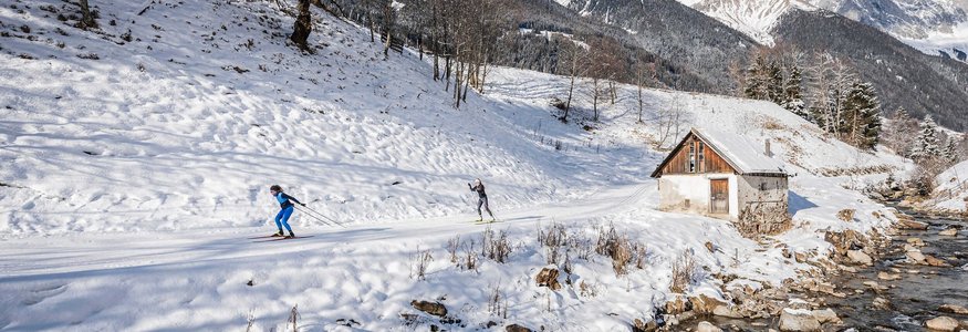 Zwei Skilangläufer auf verschneiter Bergstrecke neben Bach und Hütte