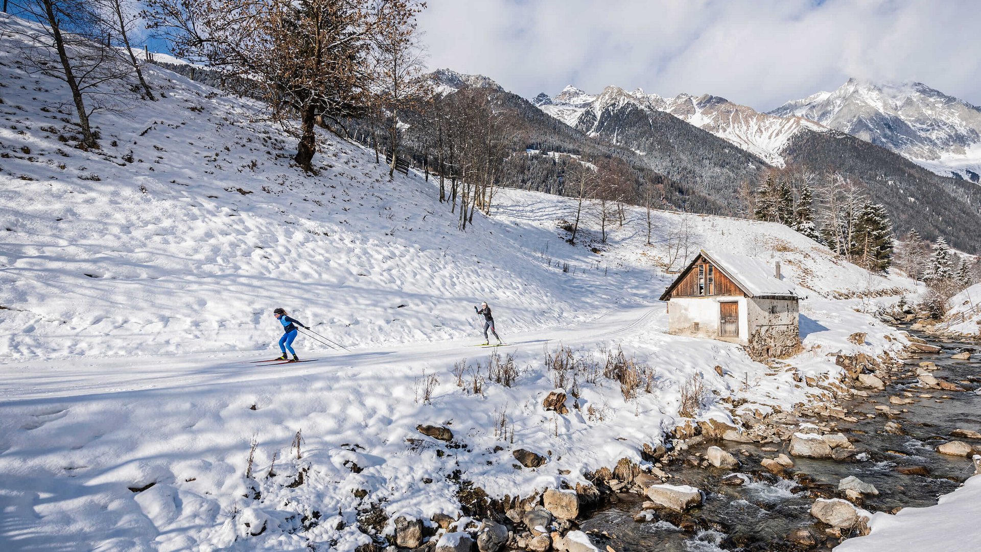 Zwei Skilangläufer auf verschneiter Bergstrecke neben Bach und Hütte